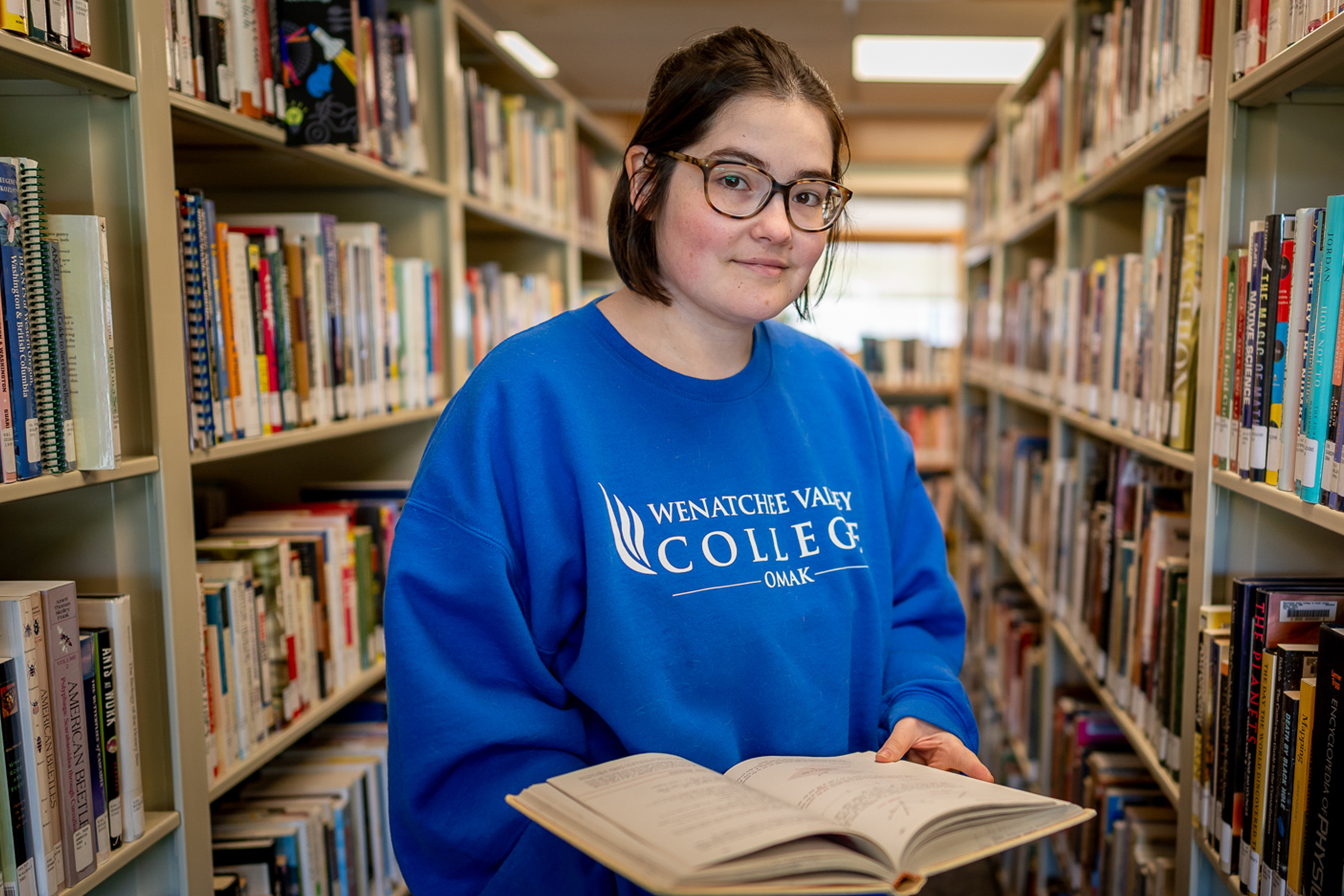 Female student with blue sweatshirt holding book in a library.