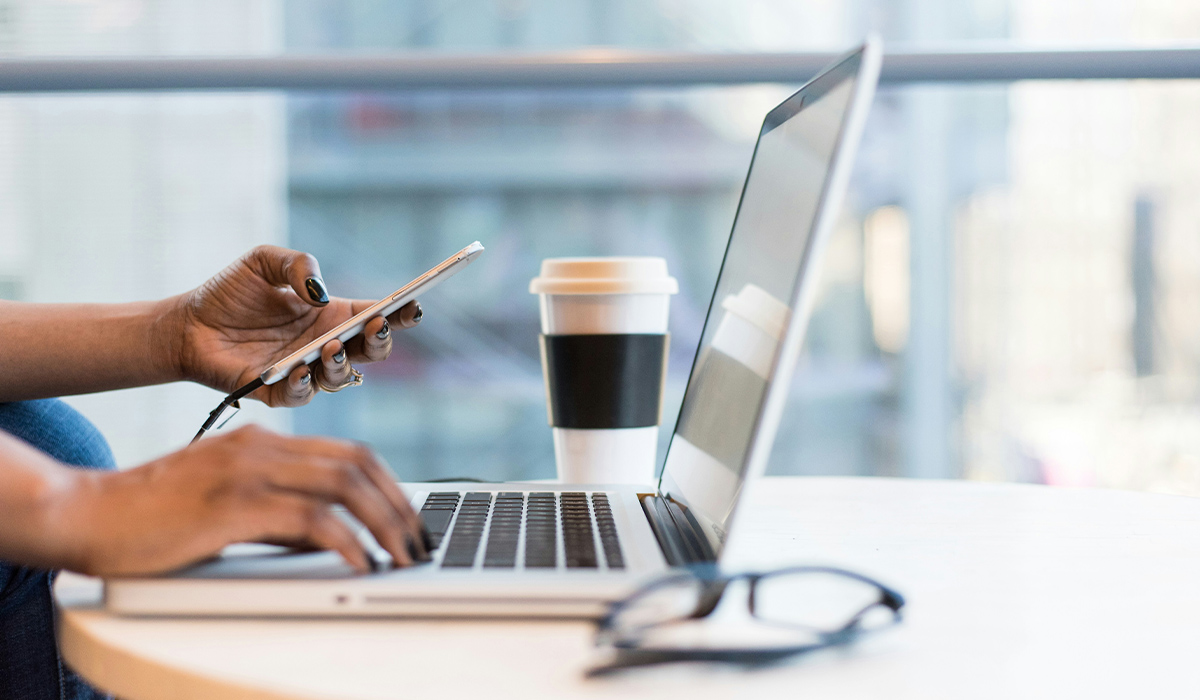 A pair of hands type on a laptop and hold a cellphone.