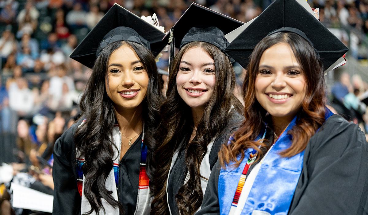Three graduates wearing black caps and gowns sit together and smile at the camera.