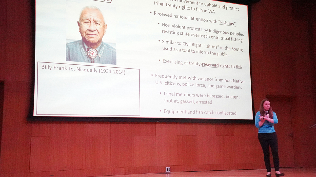Dr. Kestrel Smith stands on a stage giving a presentation about the Fish Wars.