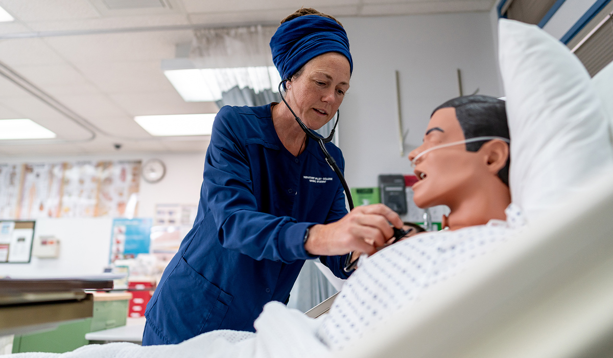 A WVC Omak nursing student holds a stethoscope over a manikin lying in a hospital bed.