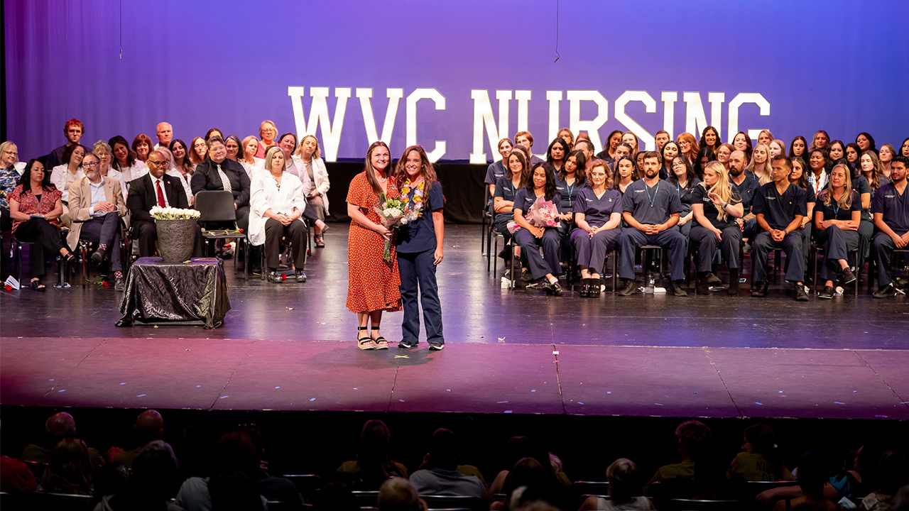 A nursing graduate and relative stand at the front of a stage with graduating nurses seated behind them.
