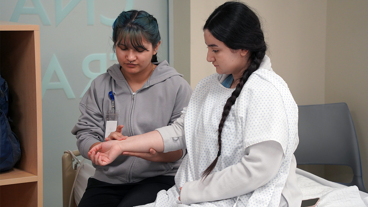 A nursing assistant student holds the arm of another nursing assistant student in order to check her pulse.