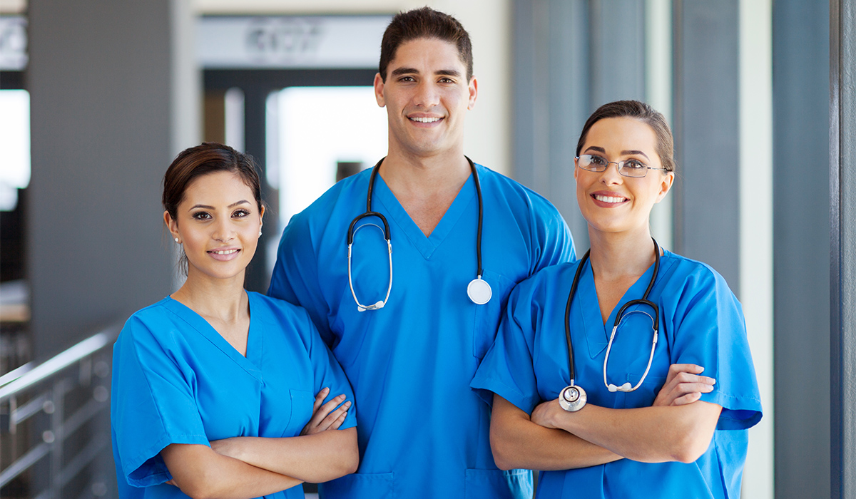 Three healthcare staff in blue scrubs stand together and smile at the camera.
