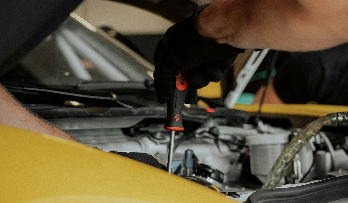 A mechanic holding a screwdriver works on a car's engine.