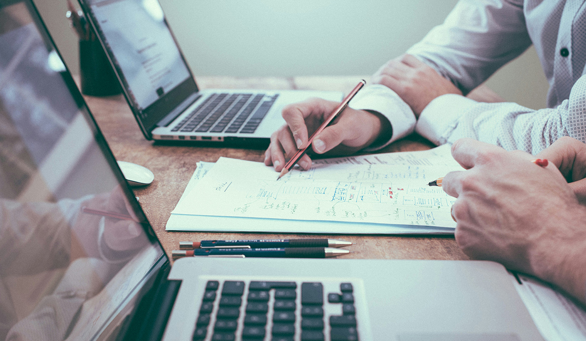 Two people sit at a table with laptops and go over a design.