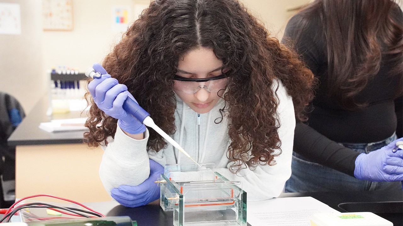A student wearing safety gear conducts an experiment with a pipette.
