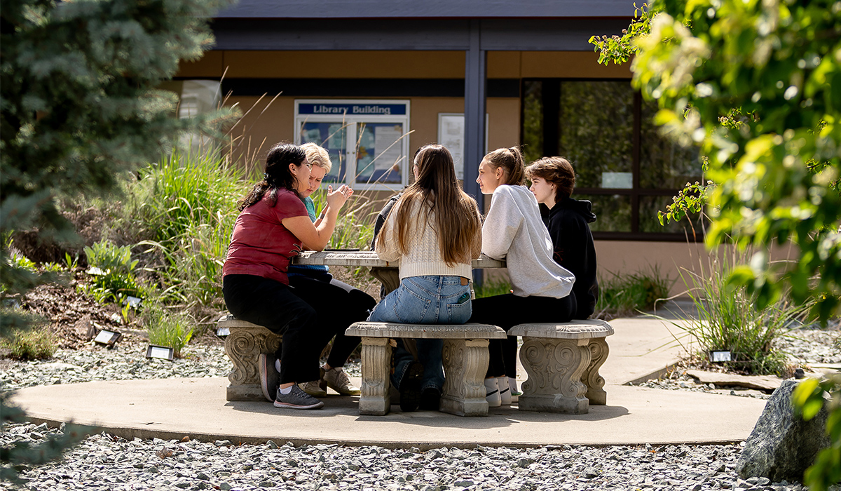 A group of students sit around a round concrete table in a native plant garden.
