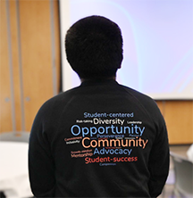 A student facing a presentation screen wears a shirt with a word cloud that has words such as "Diversity, opportunity, community, student success."